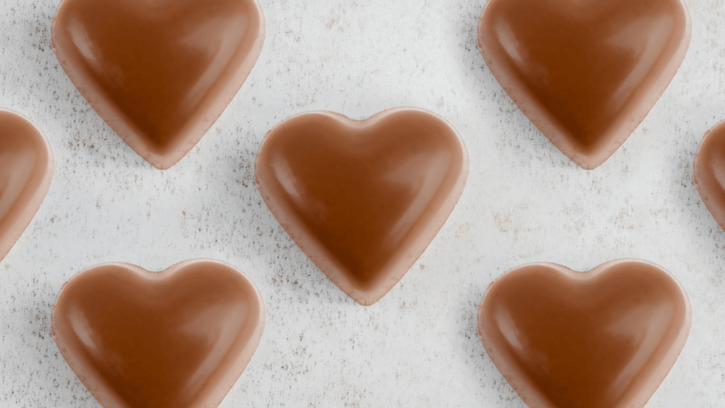 A series of glossy, milk chocolate hearts arranged in a pattern on a white marble surface, representing La Chocolatrice, a fine chocolate business, with the image conveying the brand's focus on crafted chocolate confections ideal for gifts and special occasions.