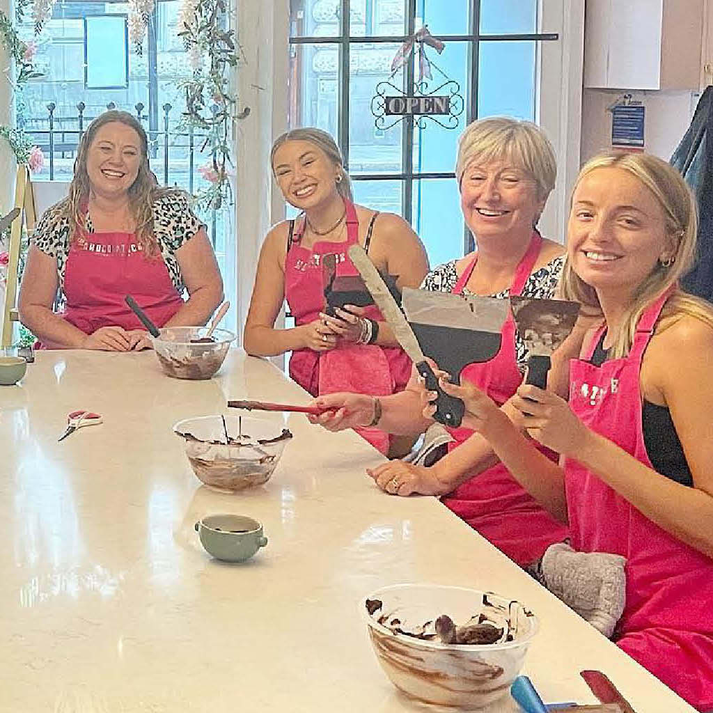A group of women in pink aprons enjoying a chocolate-making workshop at La Chocolatrice, using chocolate tools and bowls, and smiling around a table.