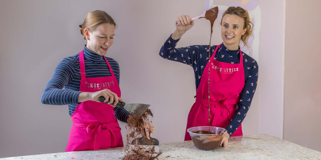 Two people enjoying a chocolate-making experience, one grating chocolate while the other pours melted chocolate from a ladle, both wearing 'La Chocolatrice' aprons, showcasing the hands-on, joyful atmosphere of the chocolate workshops provided by La Chocolatrice