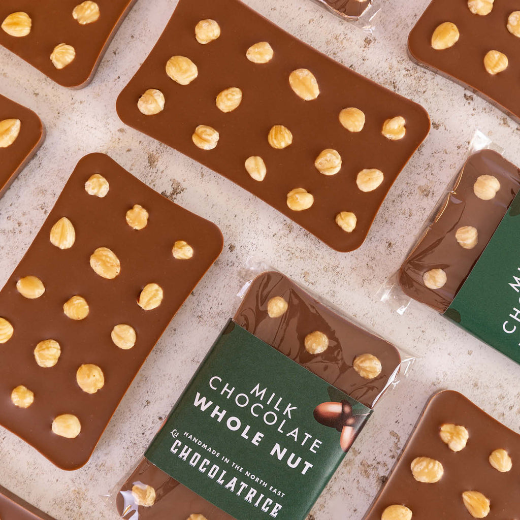 Milk chocolate bars with whole hazelnuts scattered on top, including wrapped bars labelled 'Milk Chocolate Whole Nut,' handcrafted by La Chocolatrice.