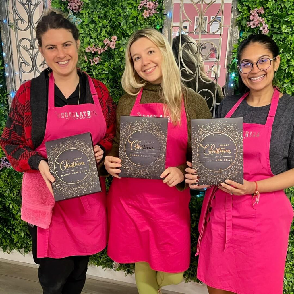 Three women proudly holding their handcrafted advent calendars, wearing pink aprons at a chocolate-making workshop.