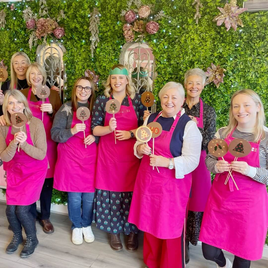 Group of women in pink aprons holding chocolate lollipops with Christmas-themed designs at a chocolate-making workshop.