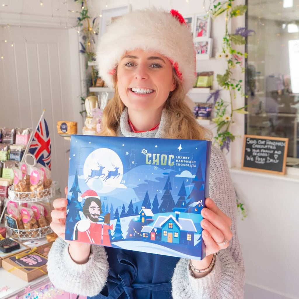 Smiling woman in Santa hat holding La Chocolatrice Christmas advent calendar inside the chocolate shop.