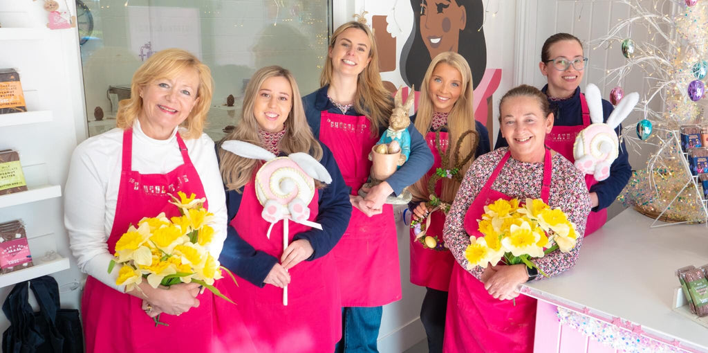 Team of chocolatiers at La Chocolatrice wearing pink aprons, holding Easter decorations and flowers inside the chocolate studio.