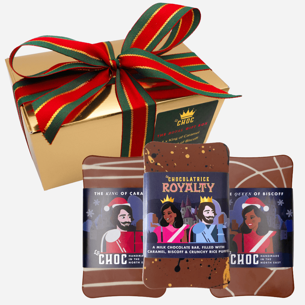 A luxurious gold gift box tied with a festive red and green striped ribbon sits behind three handmade chocolate bars. The bars are wrapped in a royal-themed Christmas design, featuring a King of Caramel bar, a Queen of Biscoff bar, and a Chocolatrice Royalty bar filled with caramel and Biscoff.