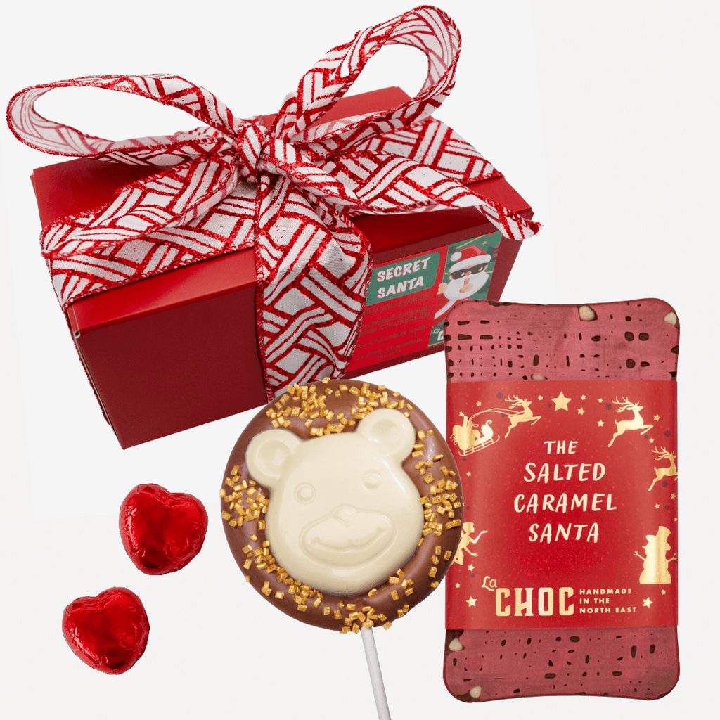 A festive red gift box with a white and red patterned ribbon sits behind a Salted Caramel Santa chocolate bar, a milk chocolate polar bear lolly with white chocolate details and gold sprinkles, and two foil-wrapped red milk chocolate hearts.